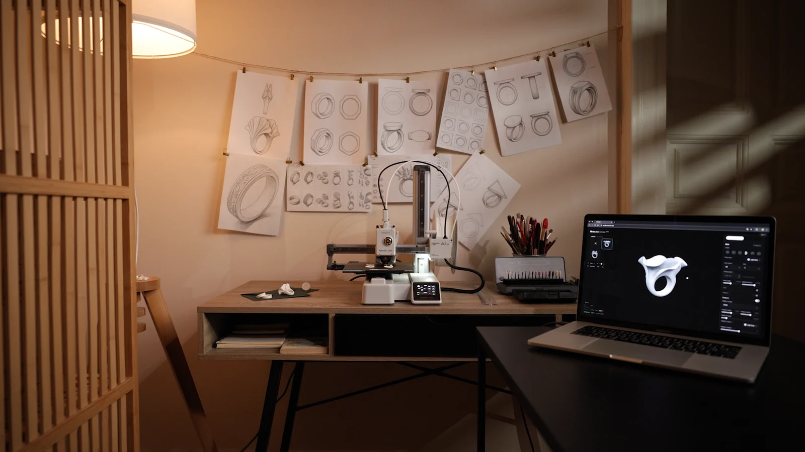 The Morcano workshop: a wall of ring sketches above a wooden desk with a Bambu Lab A1 printer, and a laptop showing a ring in Morcano Studio.